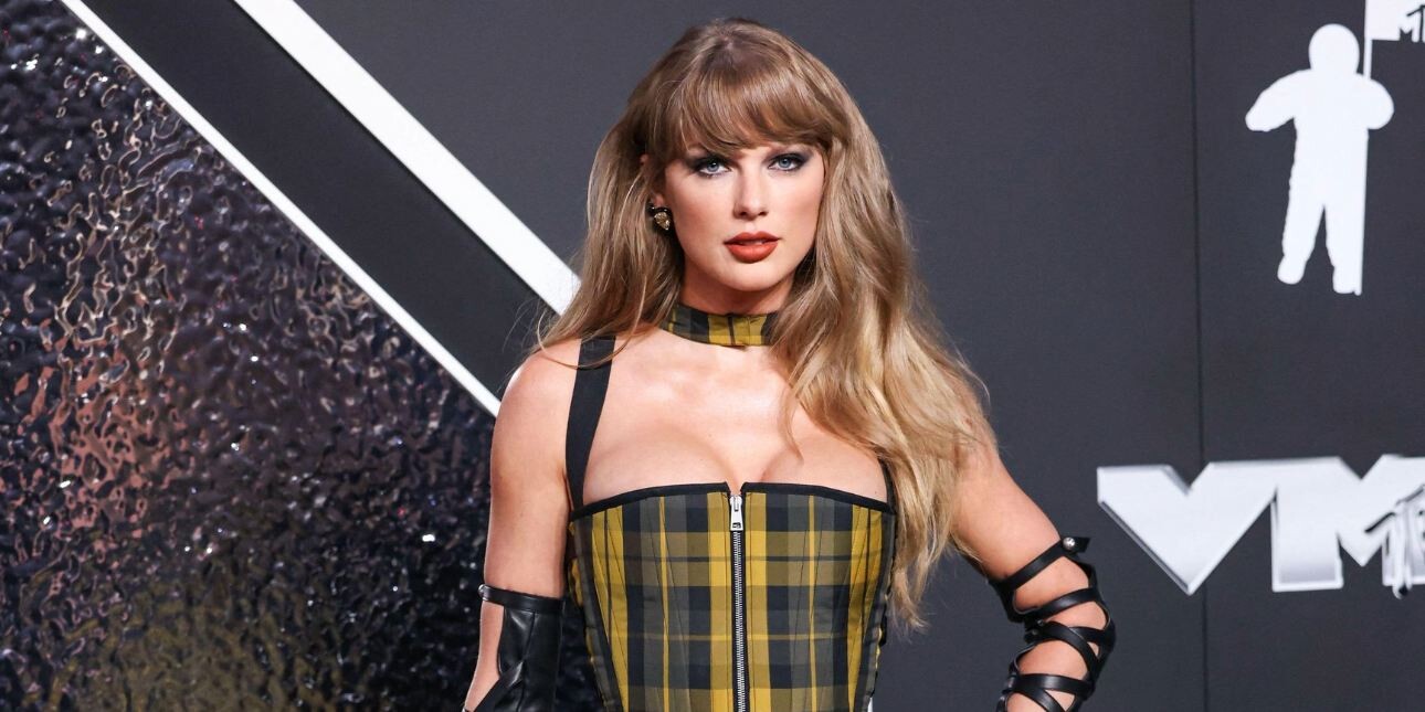 Taylor Swift poses for photographers while wearing a yellow and black check dress and black leather arm bands. The background is mostly black.